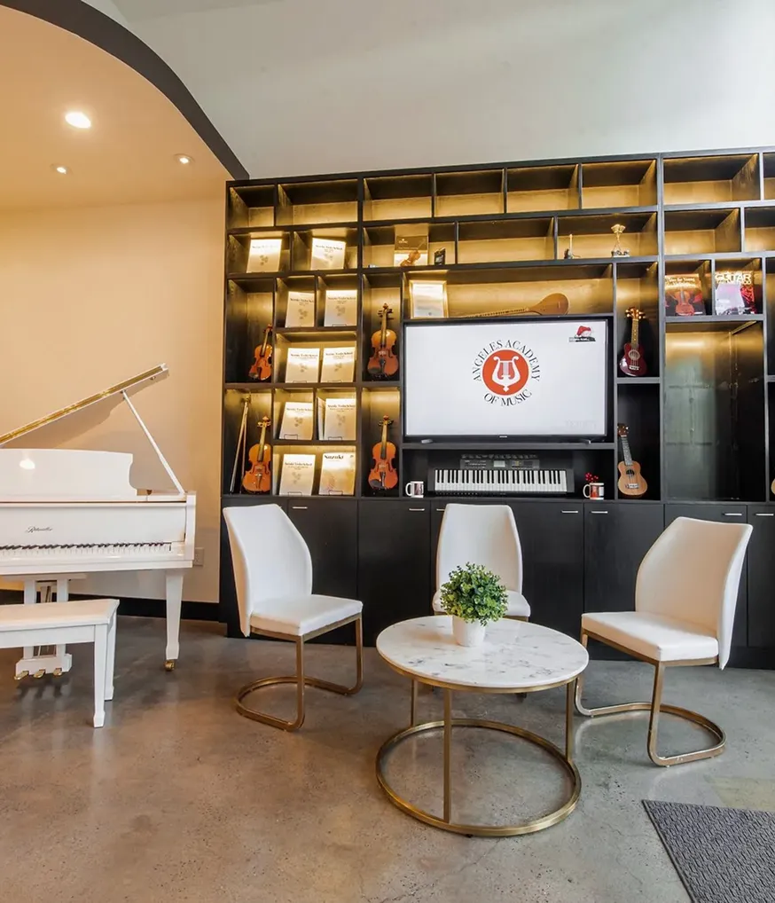 Modern music room with white grand piano and three white chairs around a circular marble table with a small green plant, set against a dark shelving unit holding violins, ukuleles, sheet music, a digital keyboard, and a TV displaying the Angeles Academy of Music logo.