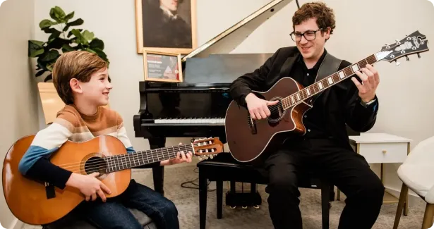 Young boy and man sitting and playing acoustic guitars together in a cozy room with a piano and framed portrait in the background.