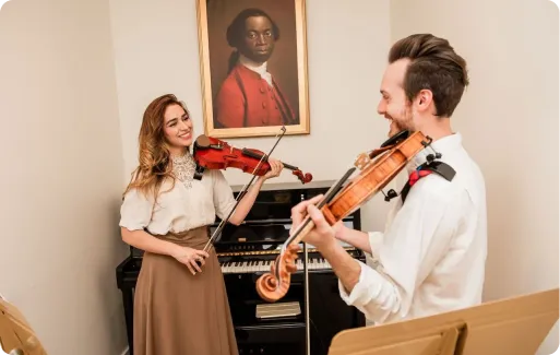 A man and a woman playing violins facing each other in a room with a piano and a framed portrait on the wall.