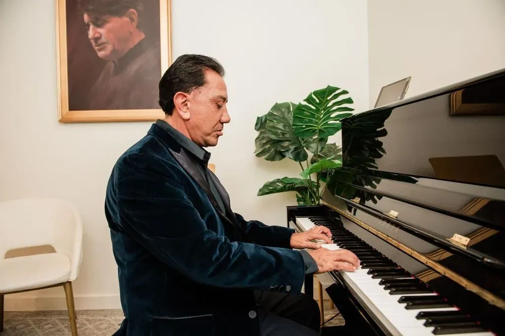 Man in a dark velvet jacket playing a black upright piano indoors, with a large green plant and a framed portrait on the wall behind him.