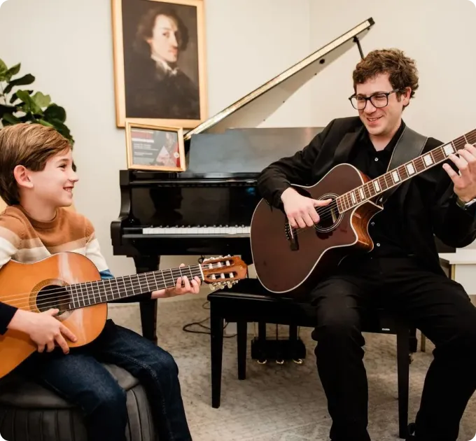 A young boy and an adult man sit facing each other, each playing an acoustic guitar, with a black grand piano and framed artwork in the background.