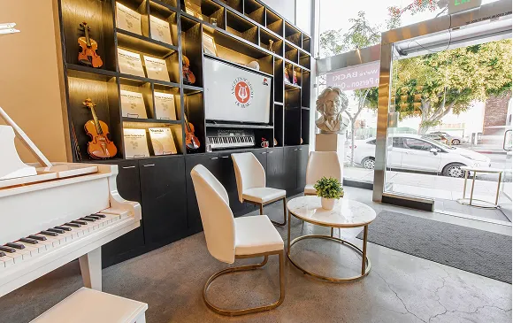 Bright music-themed lobby with white piano and chairs, round marble table with plant, violin displays, music scores on shelves, bust sculpture, and large glass window showing cars outside.