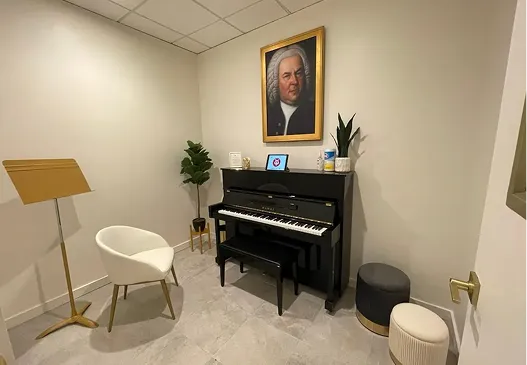 Small music room with an upright black piano, a piano bench, a white modern chair, a gold music stand, two small ottomans, a potted plant, and a framed portrait of Johann Sebastian Bach on the wall above the piano.