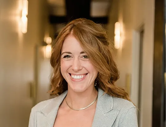 Smiling woman with wavy light brown hair wearing a gray blazer and silver necklace, standing in a softly lit hallway.