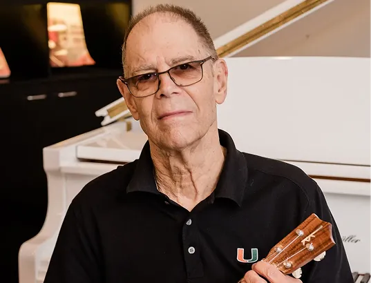 An older man wearing glasses and a black polo shirt with a University of Miami logo is holding a ukulele in front of a white grand piano.