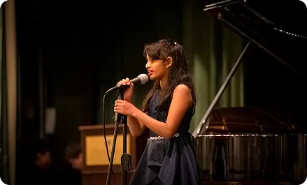 Young woman in a dark blue dress singing into a microphone on stage beside a grand piano with a dark curtain background.