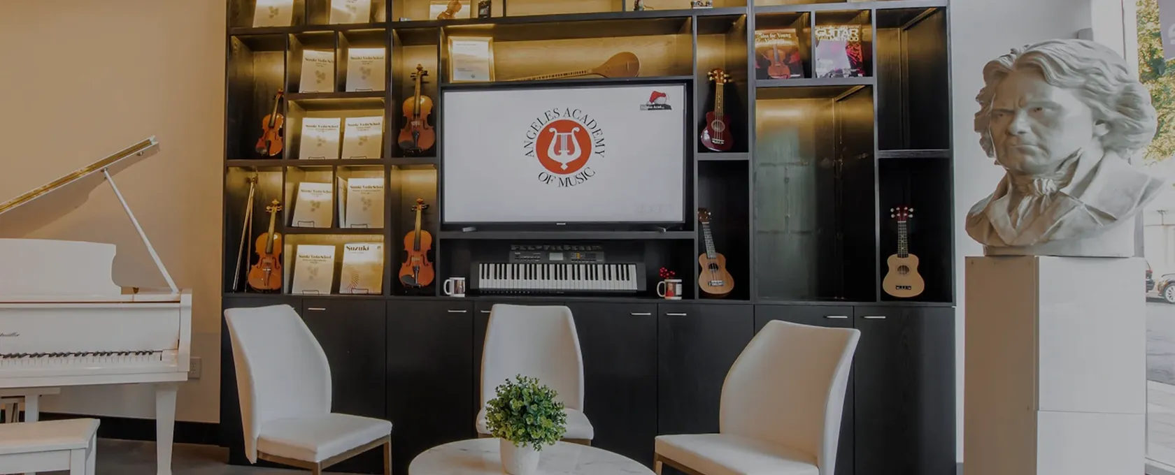 Modern music academy lobby with a white grand piano on the left, a black shelving unit filled with violins, music books, ukuleles, and a keyboard in the center, a TV screen displaying the Angeles Academy of Music logo, three white chairs around a round table with a small green plant, and a white bust sculpture on the right near a window.