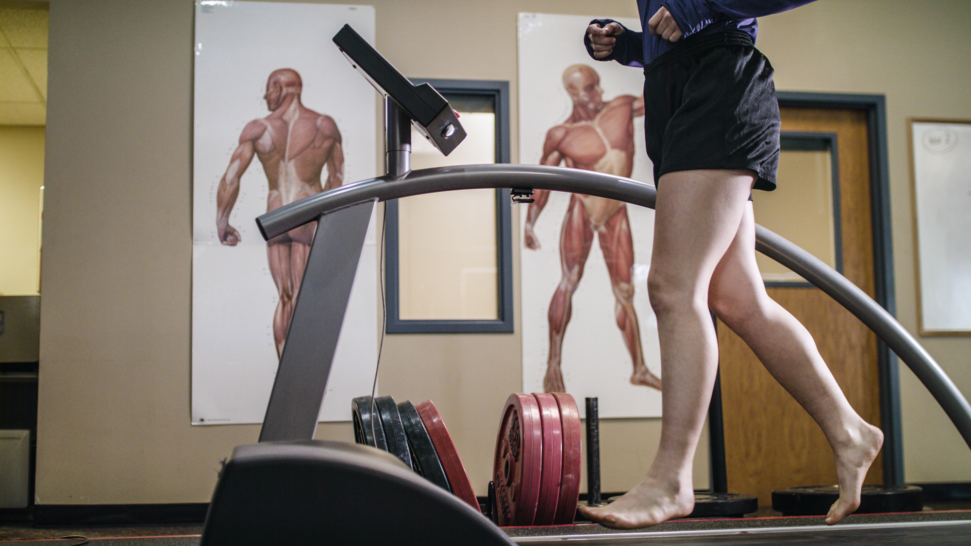 Person walking barefoot on a treadmill in a gym with anatomical muscle charts on the wall behind.