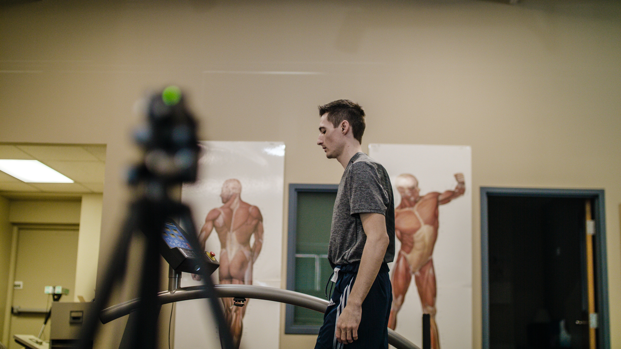 Young man walking on a treadmill in a laboratory with muscular anatomy posters on the wall behind him.