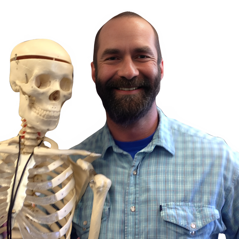 Smiling man with a beard wearing a blue plaid shirt standing next to a human skeleton model.
