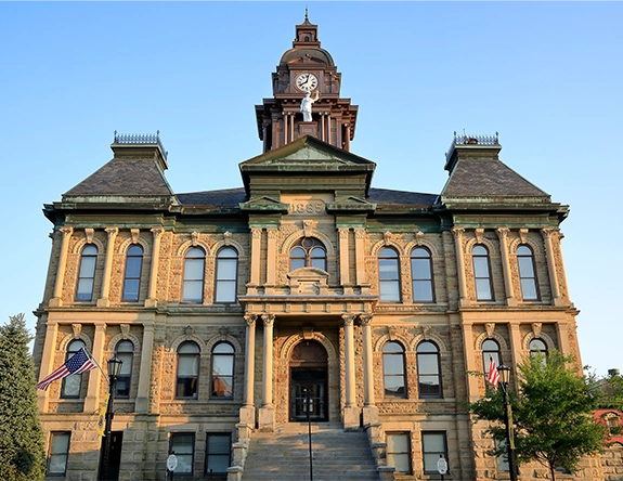 Historic stone courthouse building with a clock tower and American flags against a clear blue sky.
