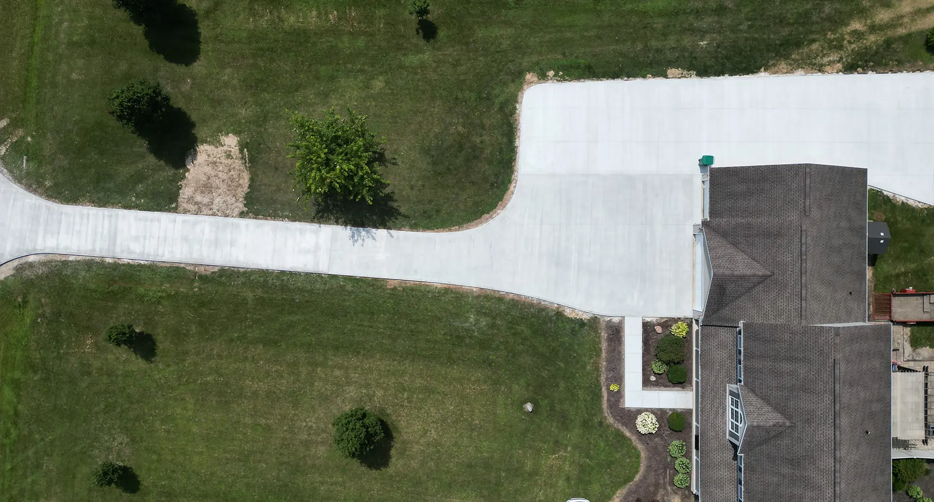 Aerial view of a residential house with a large concrete driveway and green grassy lawn with scattered bushes.