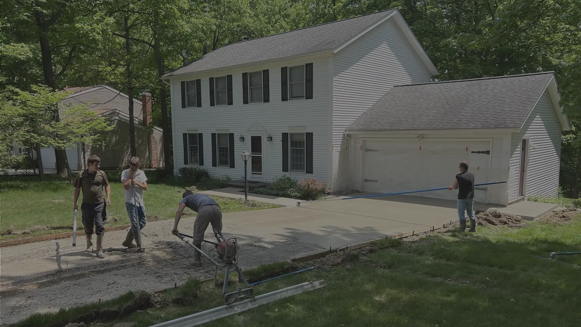 Workers smoothing and finishing a concrete driveway in front of a white two-story house and garage.