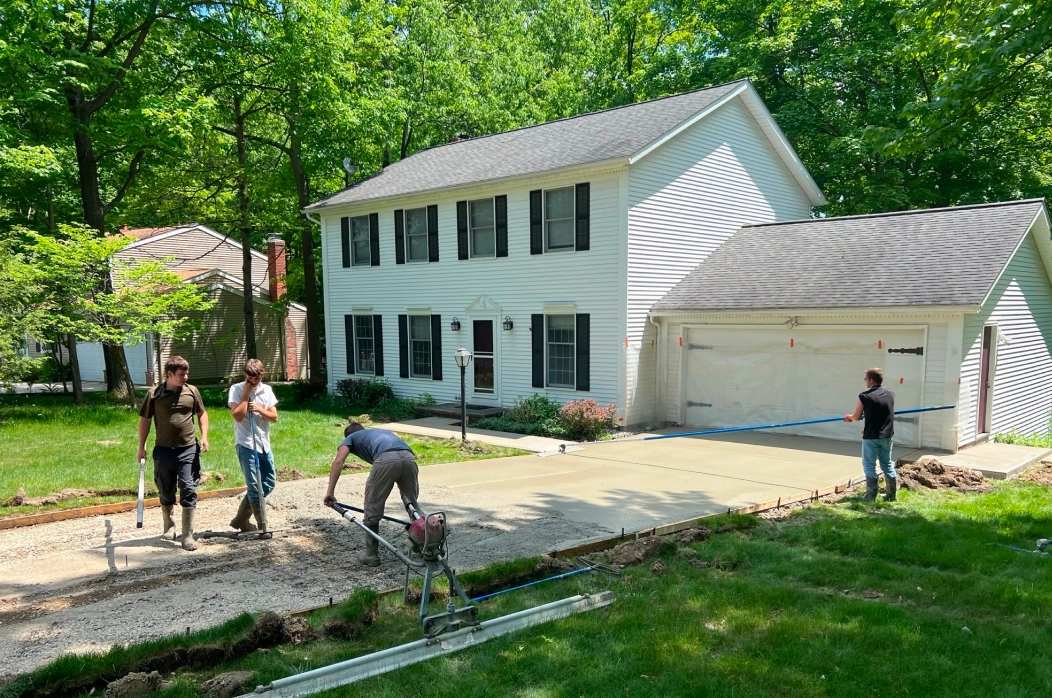 Workers spreading and smoothing freshly poured concrete for a driveway in front of a white two-story house with black shutters.