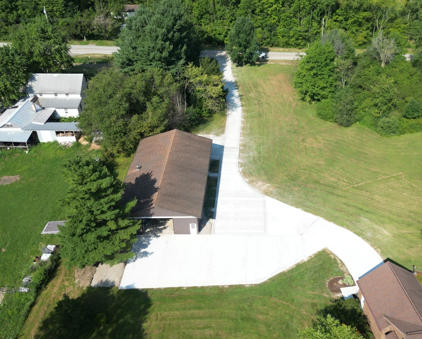 Aerial view of a residential property with a long, newly paved concrete driveway leading to a detached garage surrounded by green trees and grass.