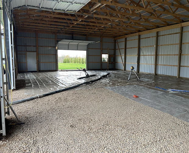 Interior of a large metal-framed barn with gravel floor and plastic sheeting prepared for concrete pouring, with construction tools inside.