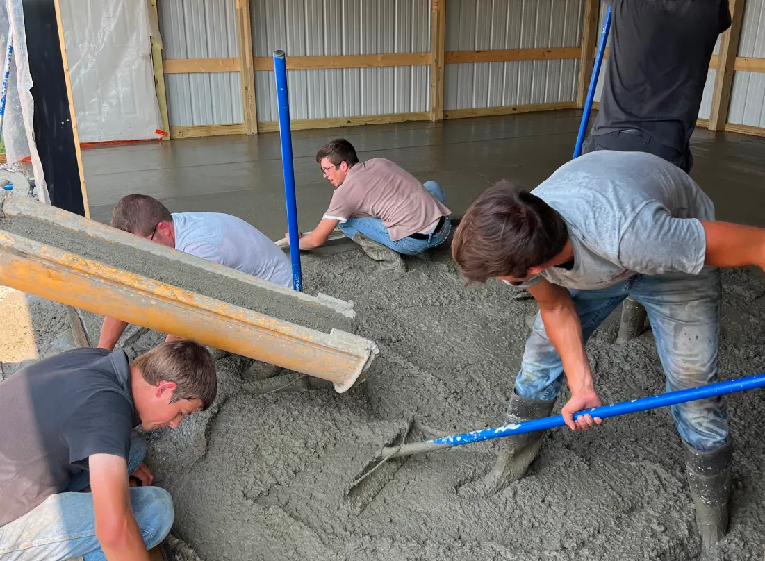 Workers spreading and smoothing freshly poured concrete for a driveway in front of a white two-story house with black shutters.