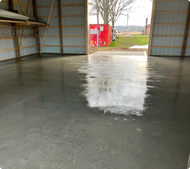 Smooth, freshly poured concrete floor inside a large metal garage with an open door showing red containers and a tree outside.