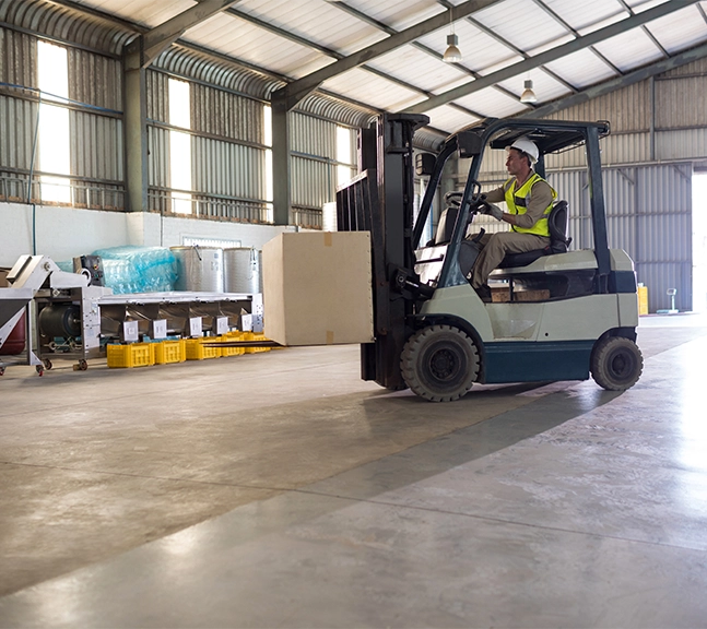 Worker wearing safety helmet and vest operating a forklift carrying a large cardboard box inside a spacious warehouse.