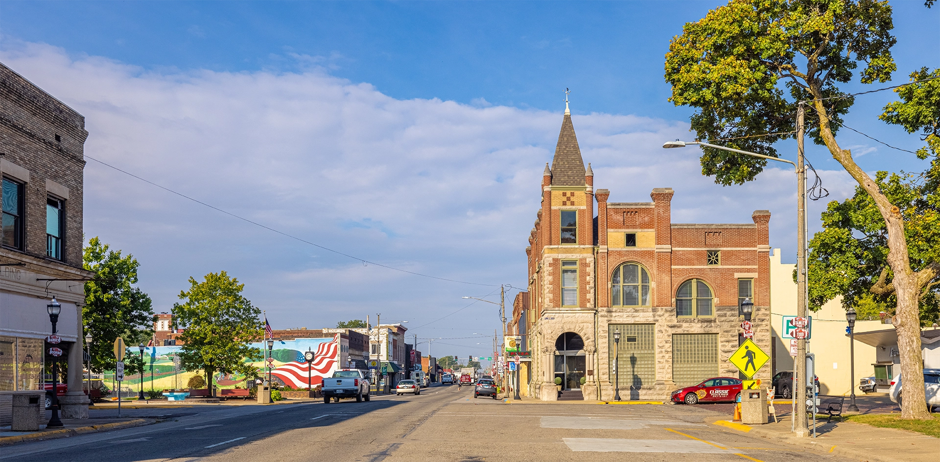 Small town street with historic brick building, cars, trees, and a mural of an American flag.