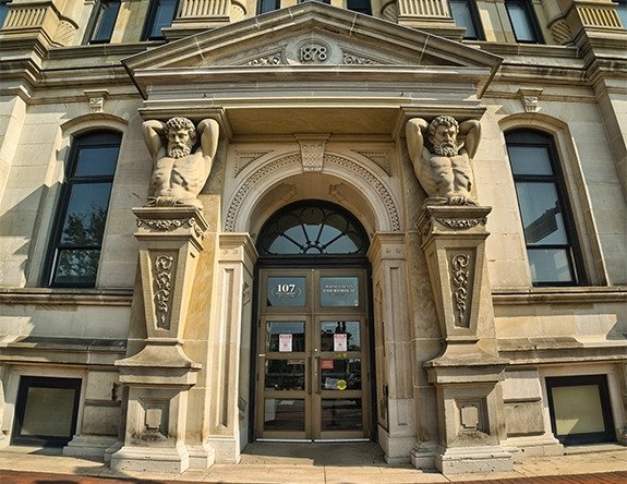 Stone courthouse entrance with ornate pillars featuring male statues and the year 1878 above the door.