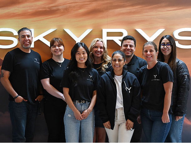 A group photo of the people team in front of the Skyryse logo on the wall