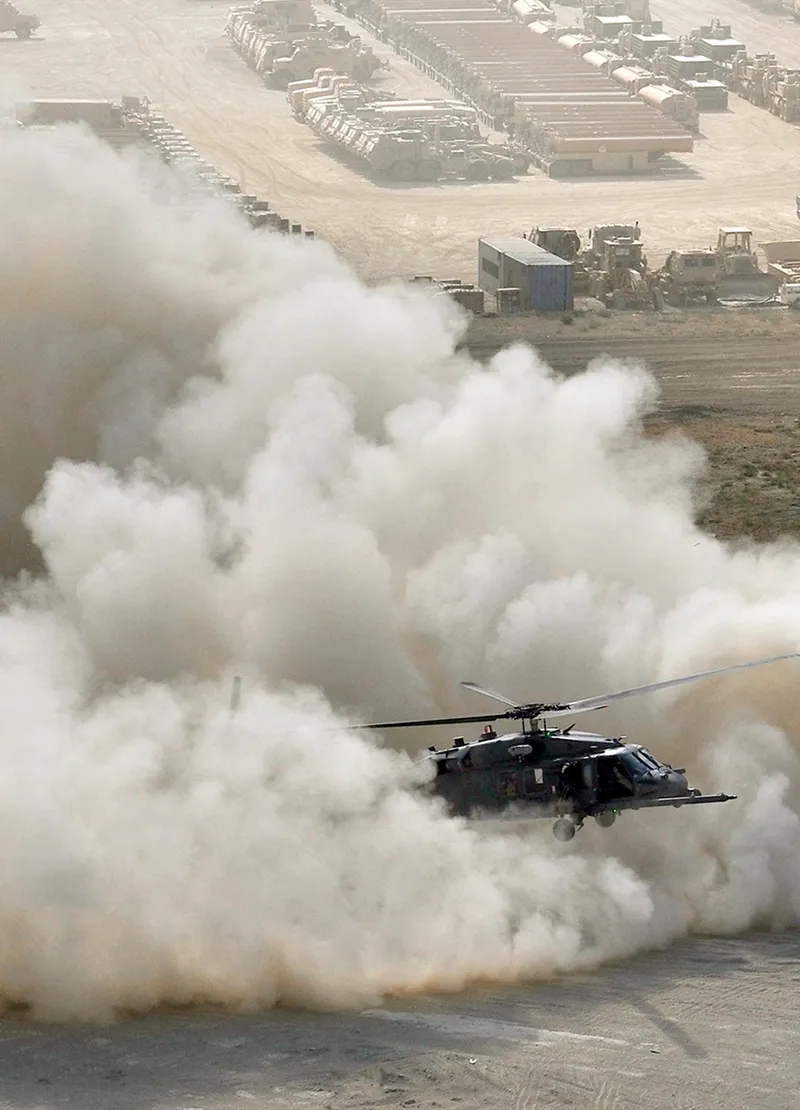 Military helicopter flying low over dusty terrain with clouds of dust and military vehicles in the background.