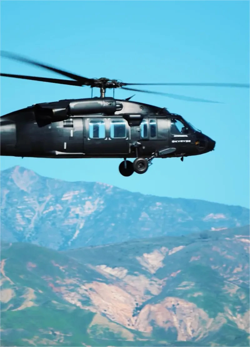 Black helicopter flying over mountainous terrain with clear blue sky.