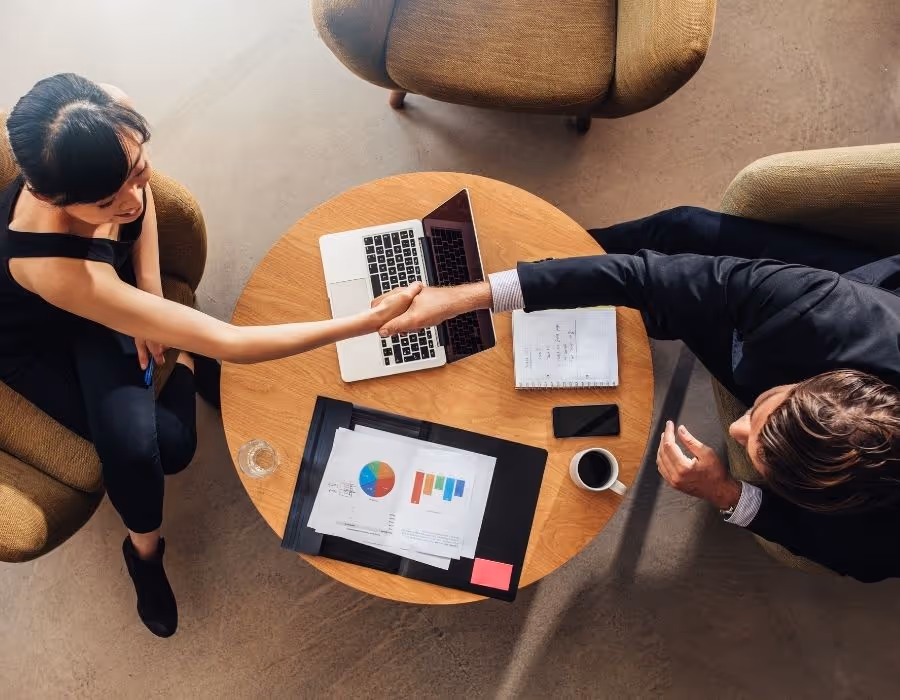 Two business professionals shaking hands over a table with a laptop and a printed report showing charts and graphs