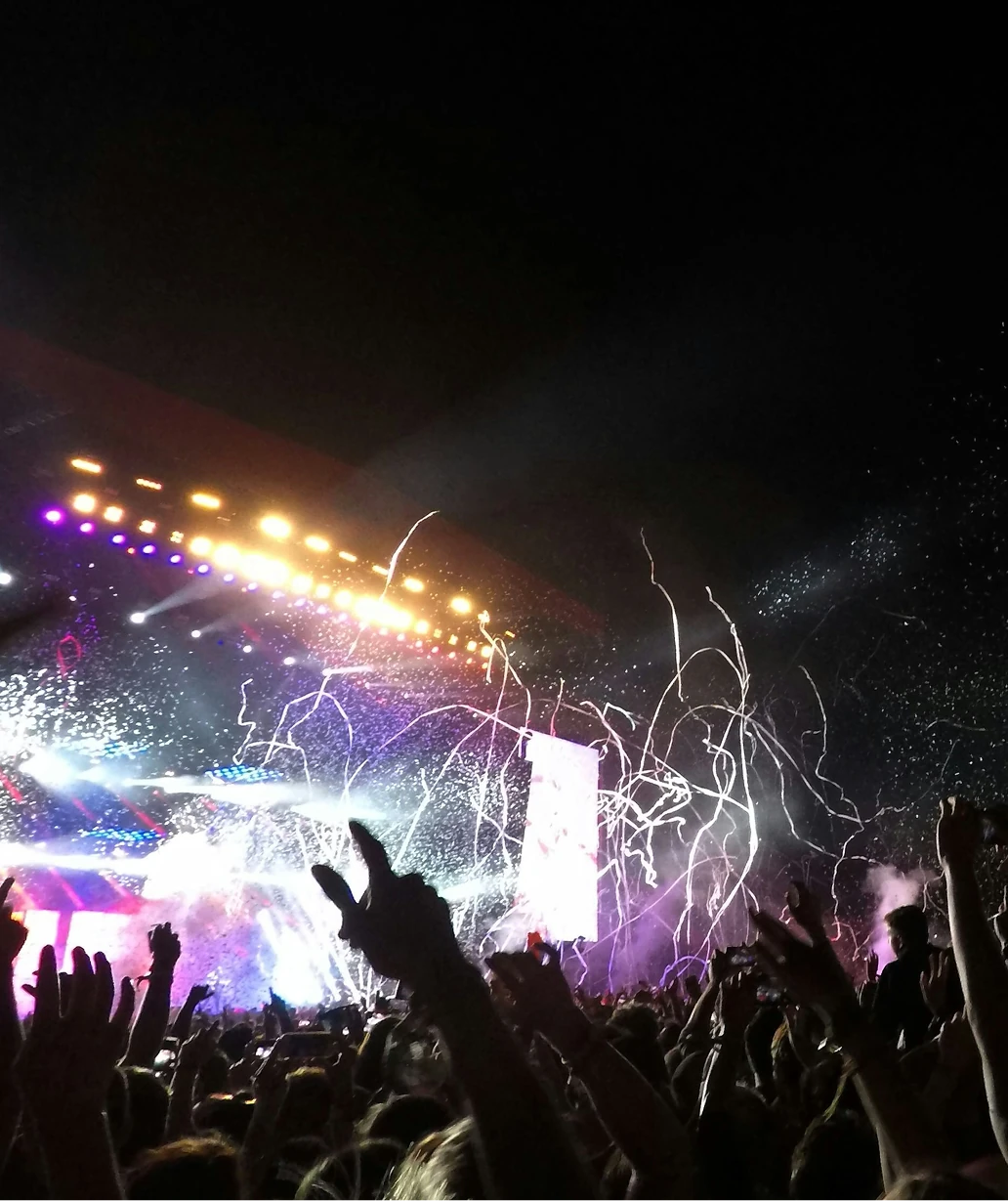 Crowd with raised hands at a nighttime concert, with stage lights and streamers filling the air
