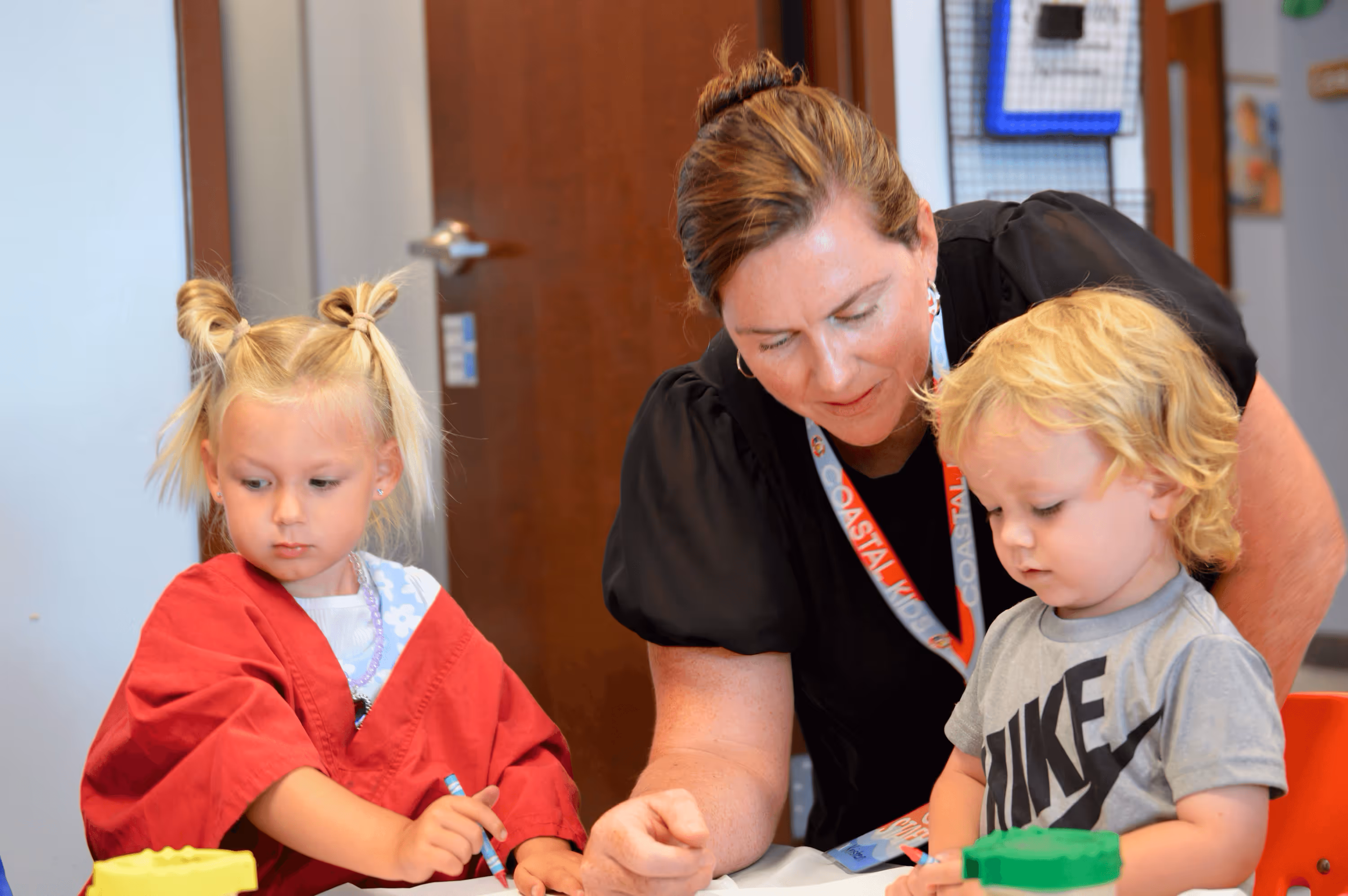 A woman in a black shirt and lanyard helps two young children with coloring activities at a table.