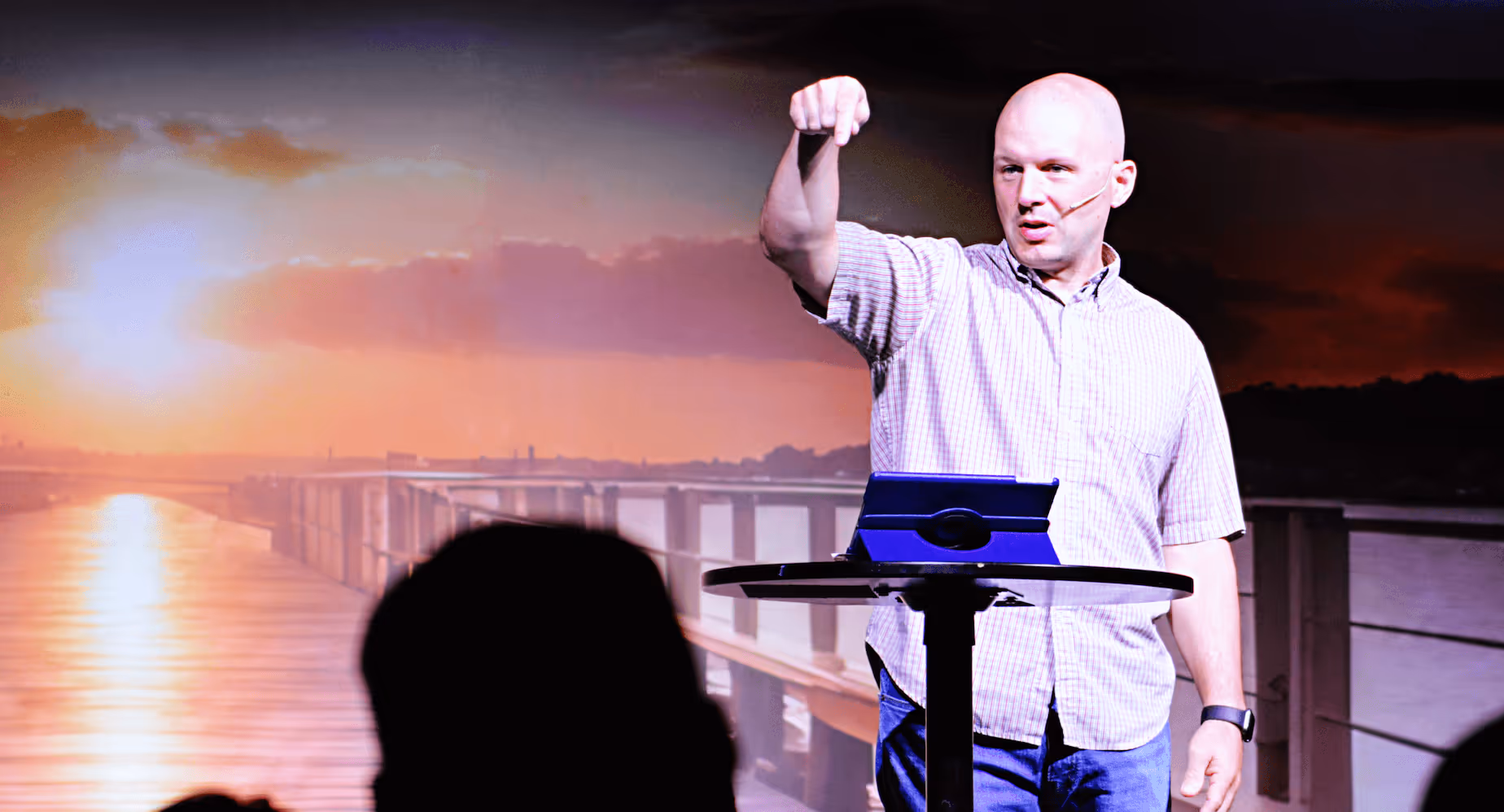 Man with a microphone headset giving a presentation on stage in front of a sunset bridge backdrop.
