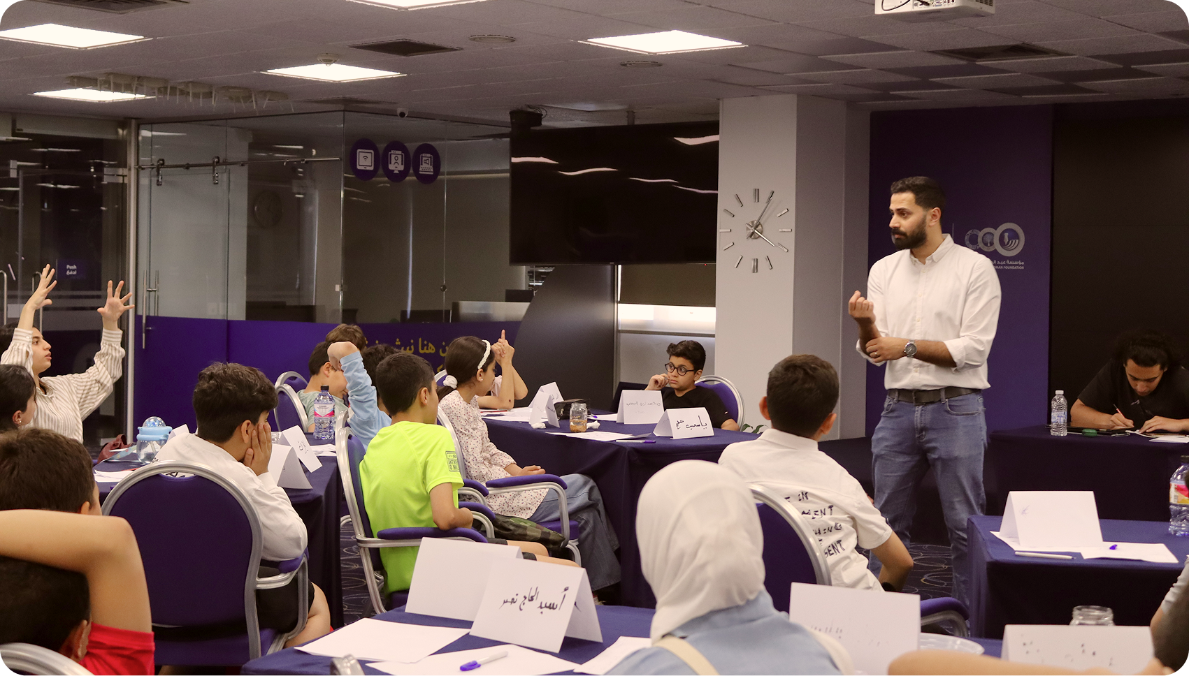Man teaching children seated at tables in a classroom setting, some kids raising hands.