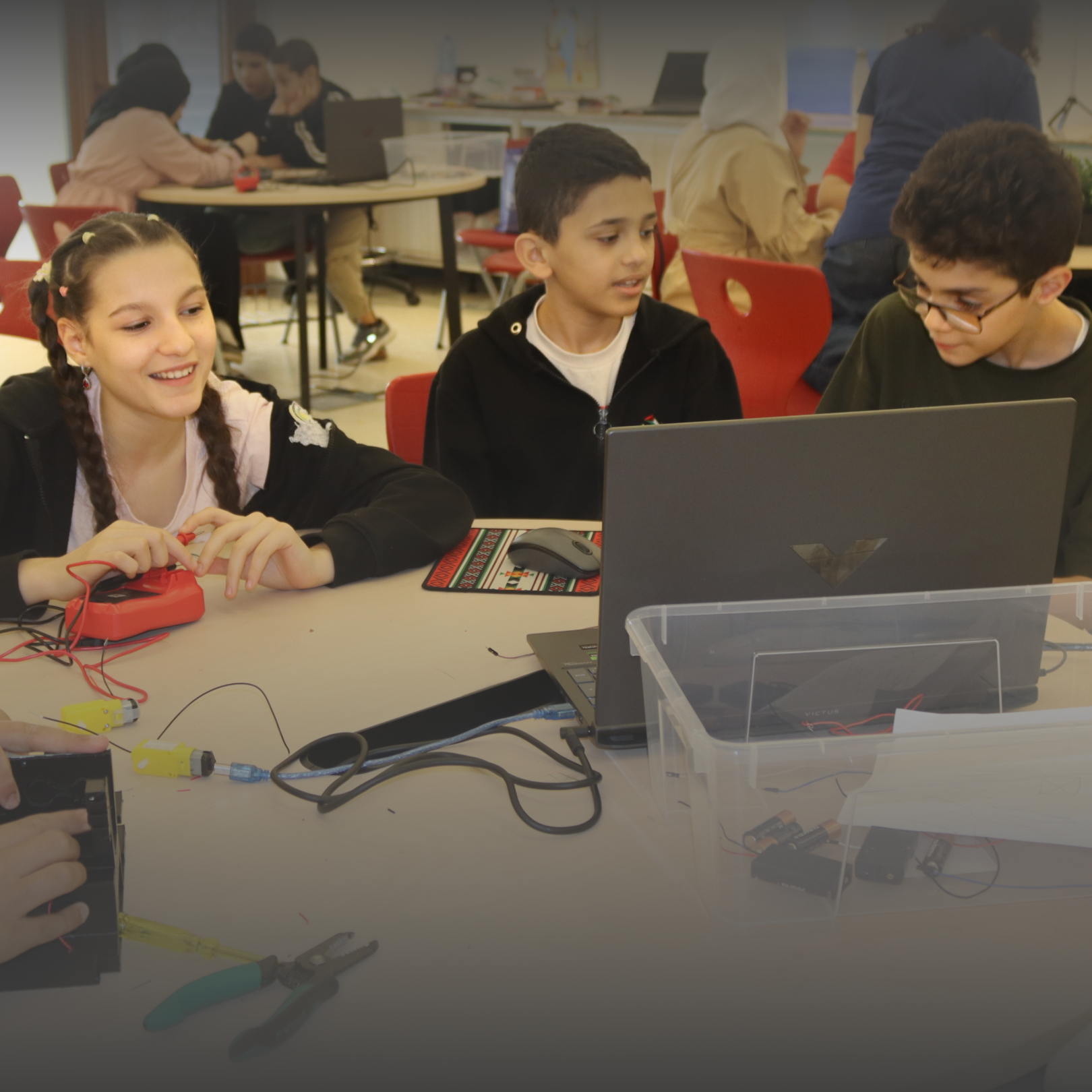 Three children collaborate on a laptop and electronic components in a classroom setting.