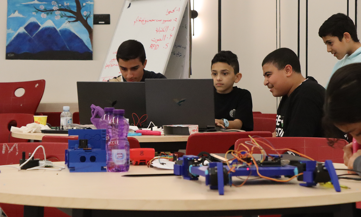 Group of boys working on laptops with electronic and robotic components on tables in a classroom.