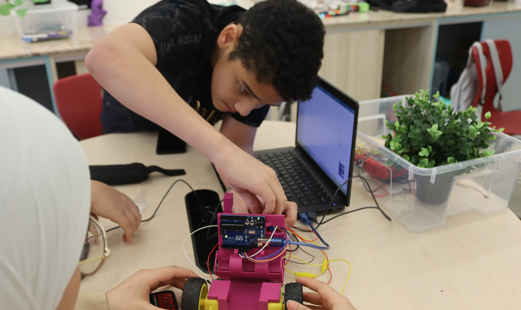 Two children working on a pink robot car with wires and an Arduino board next to a laptop.