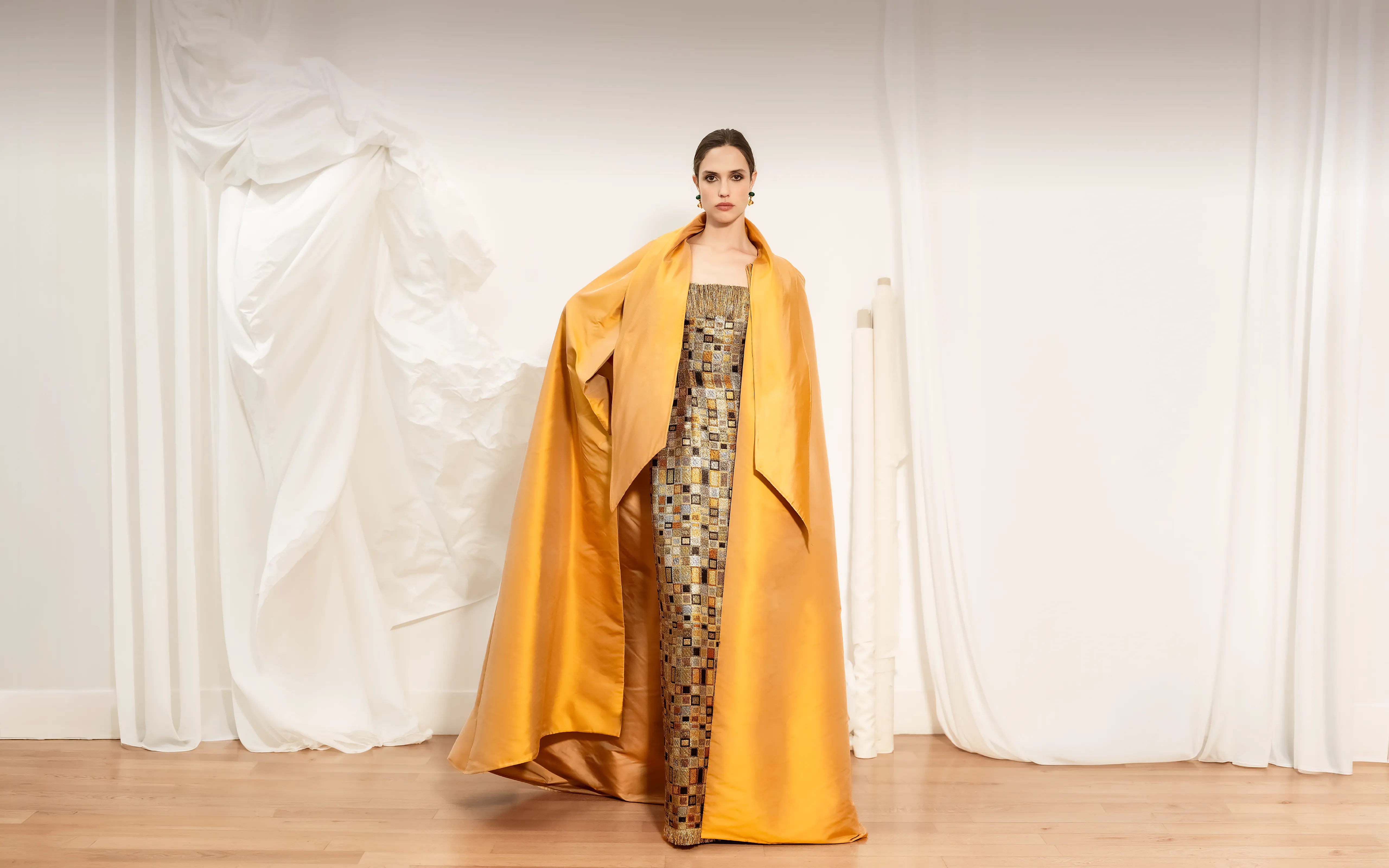 Woman standing on wooden floor in a minimalist room with white draped curtains, wearing a strapless dress with a mosaic pattern and a large golden satin cape draped over her shoulders.