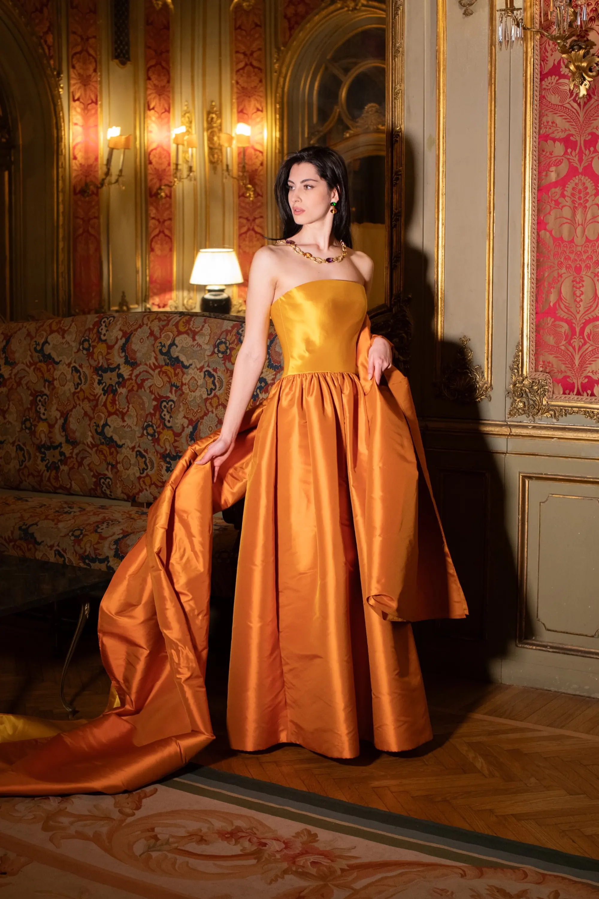 Model wearing a strapless orange satin gown with a full skirt and long train in an ornate historic interior.