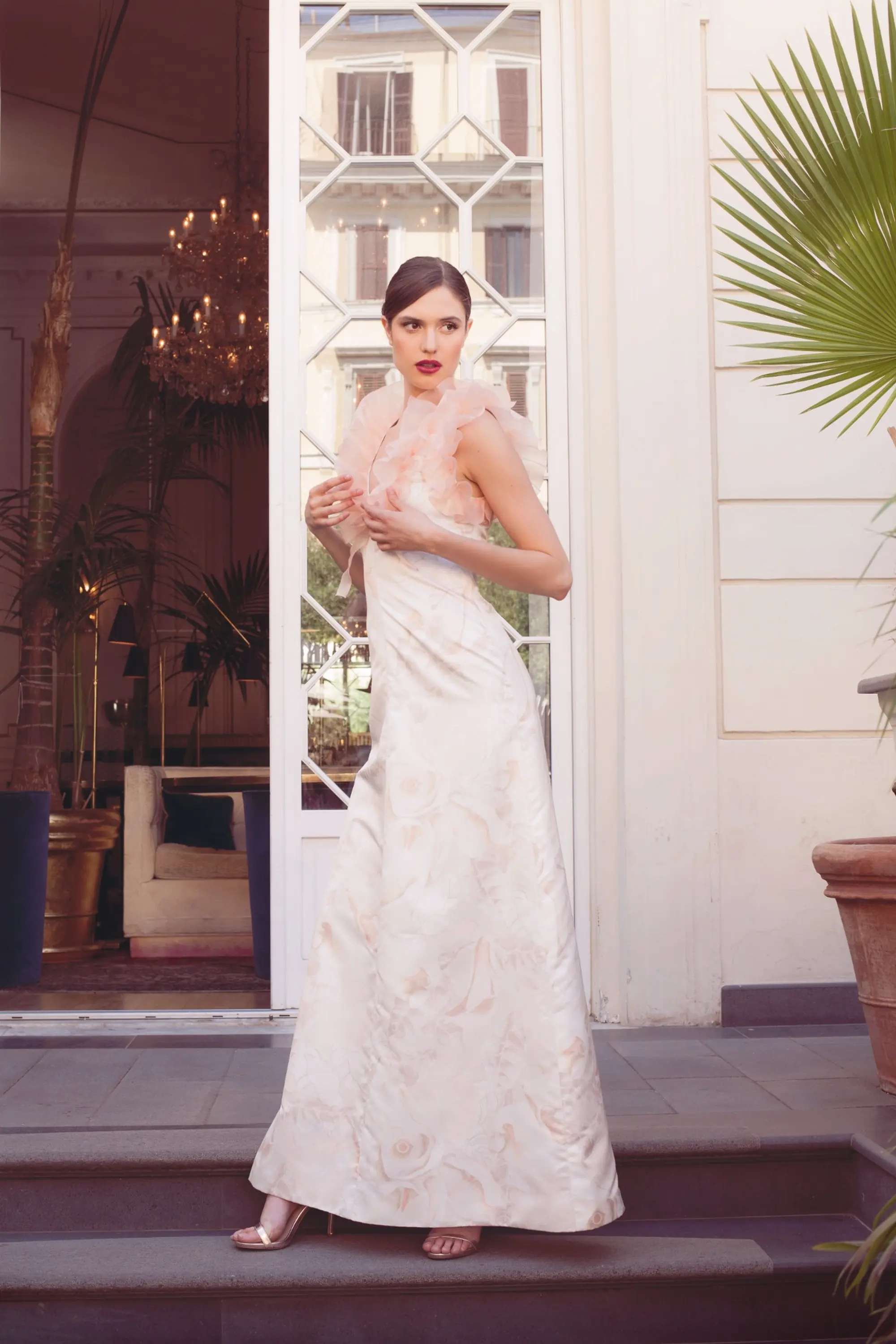 Model wearing a long ivory gown with soft pink ruffle detailing at the shoulder, standing on the steps of an elegant entrance.