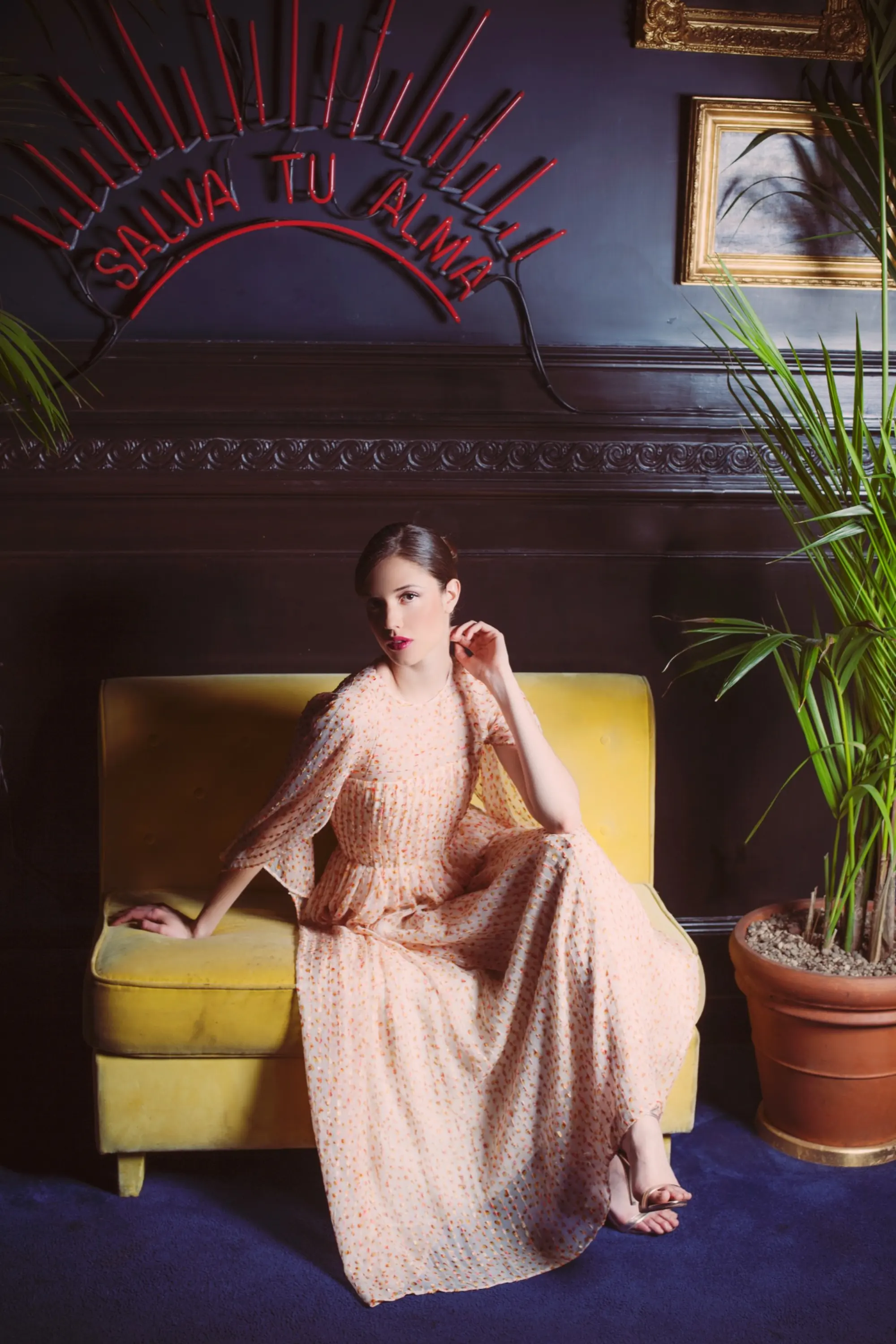 Model wearing a long pale dress with a delicate orange dotted pattern, seated on a yellow armchair in an elegant interior.