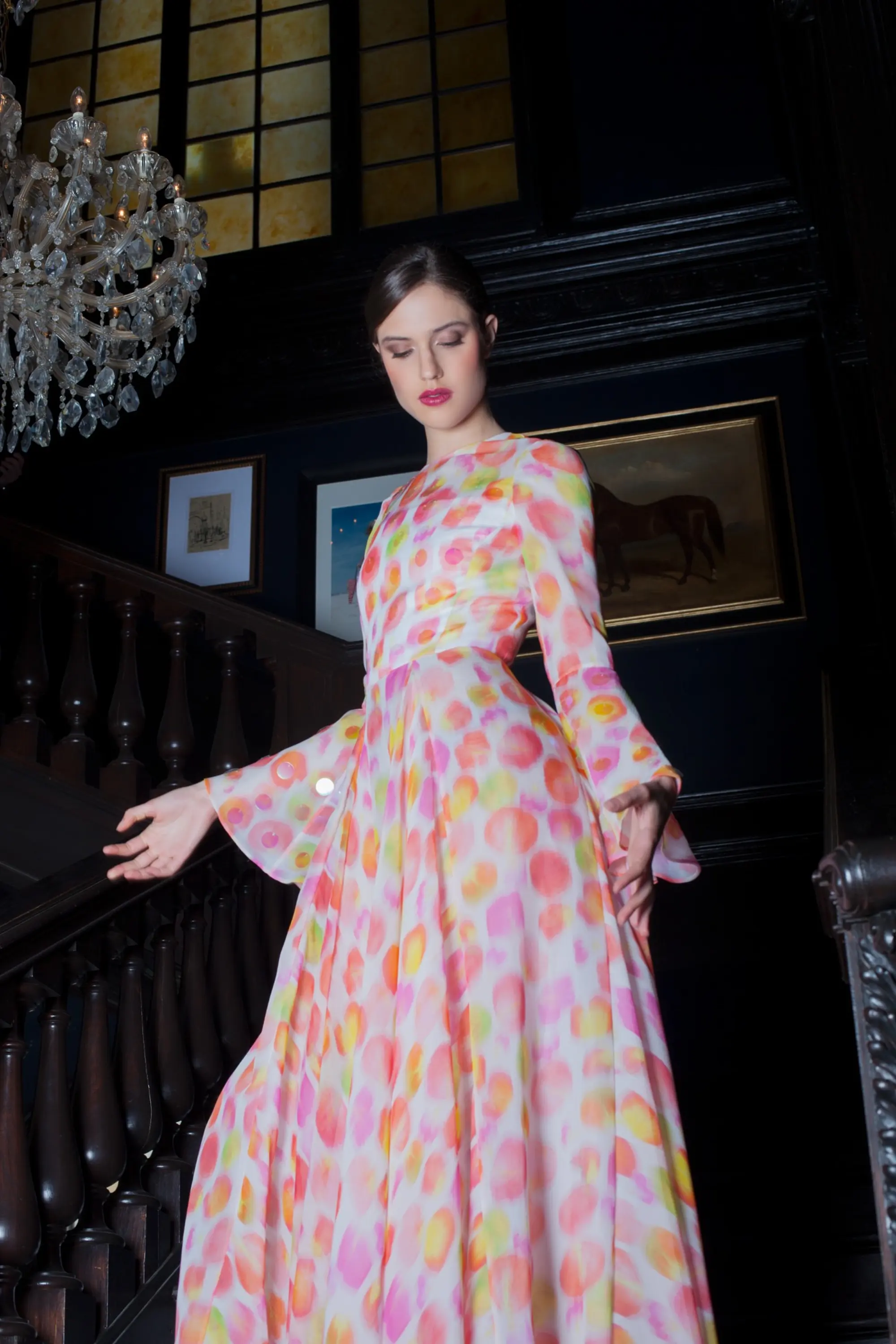 Model wearing a long white dress with a colorful pink, orange and green circular pattern, standing beside a dark wooden staircase beneath a crystal chandelier.