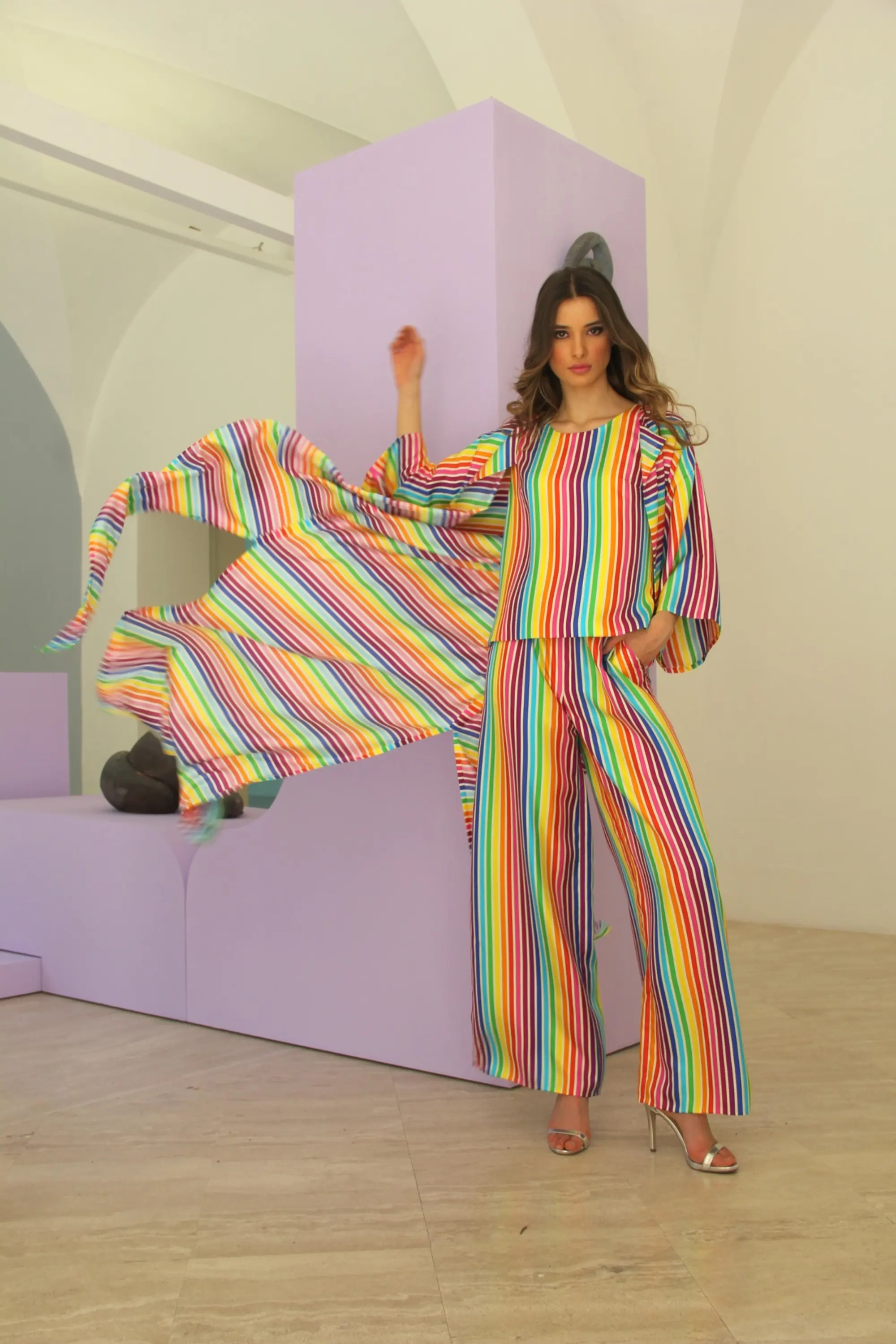 Model wearing a rainbow-striped ensemble with wide-leg trousers and a flowing cape, standing in a minimalist interior with pastel lavender geometric structures.
