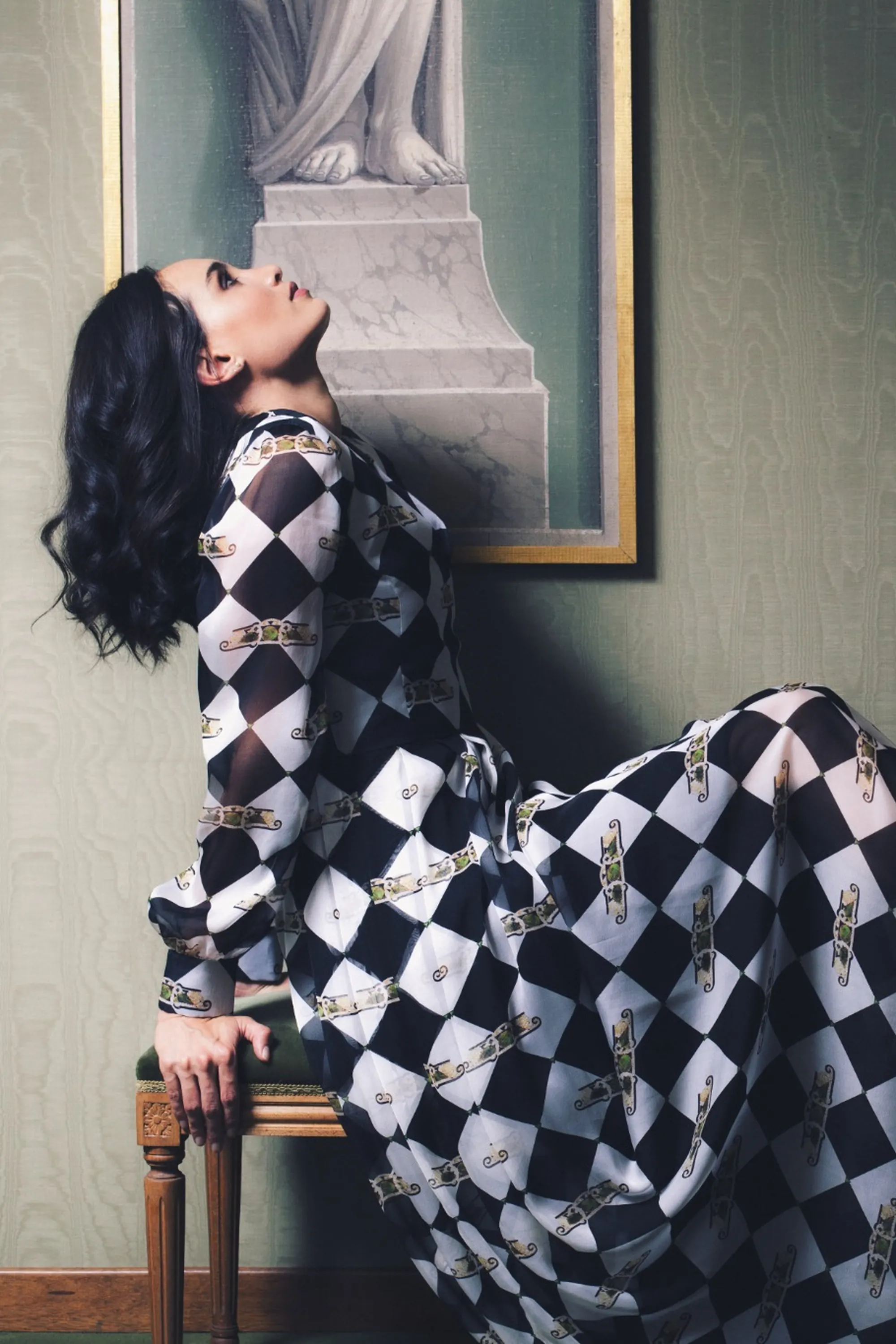 Model wearing a black-and-white diamond-patterned dress, posed elegantly between two green upholstered benches in a refined interior.