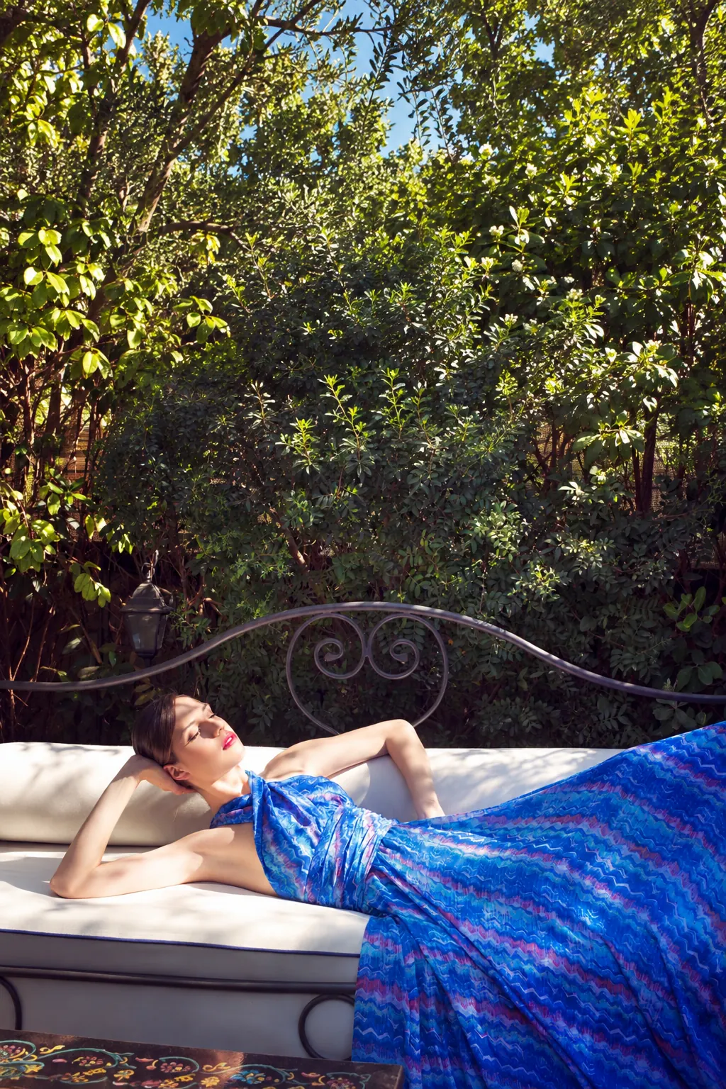 Model reclining outdoors in a flowing blue patterned gown on a cushioned daybed surrounded by greenery.