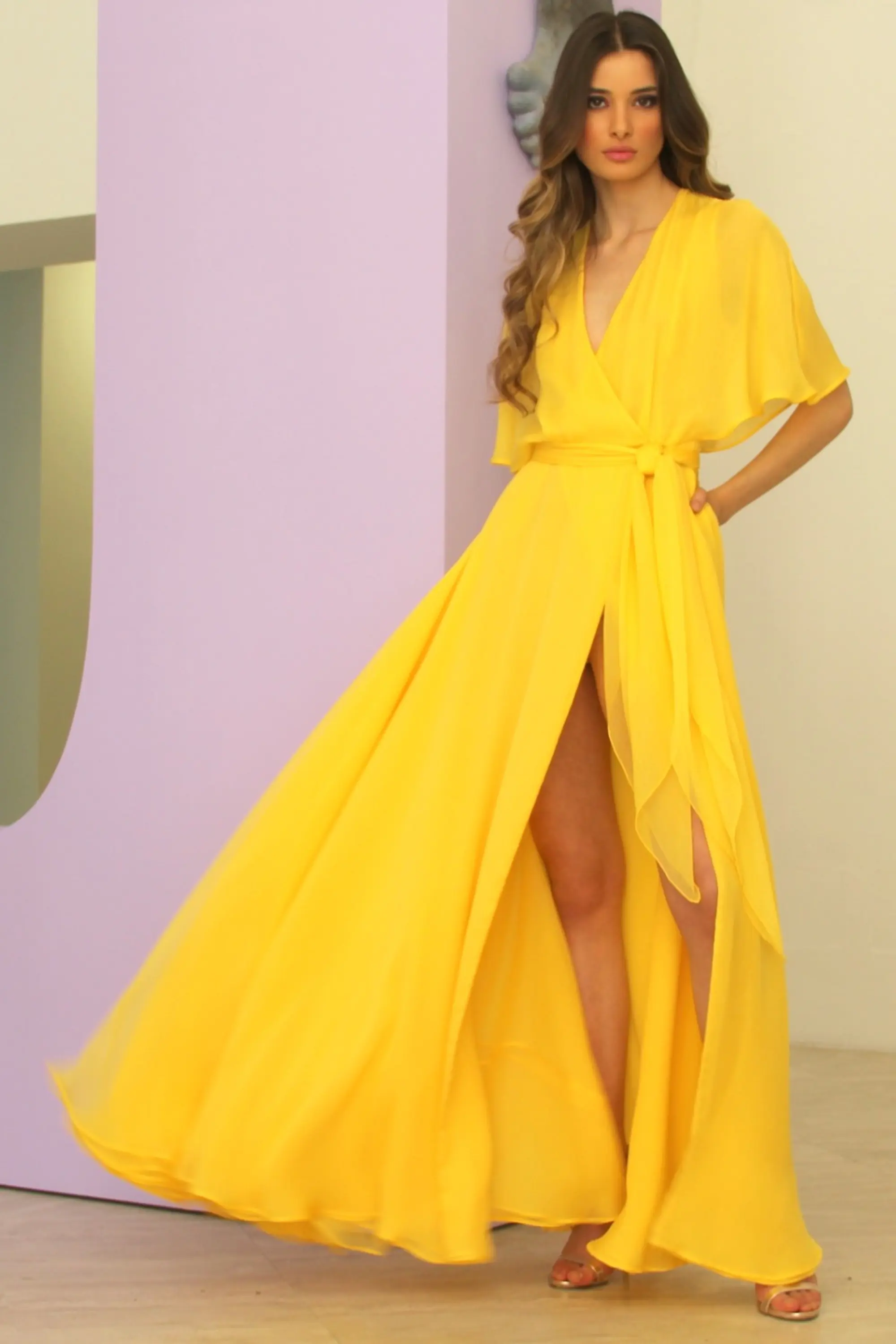 Model wearing a flowing yellow floor-length dress with a deep V-neck and waist tie, standing in a minimalist pastel interior.