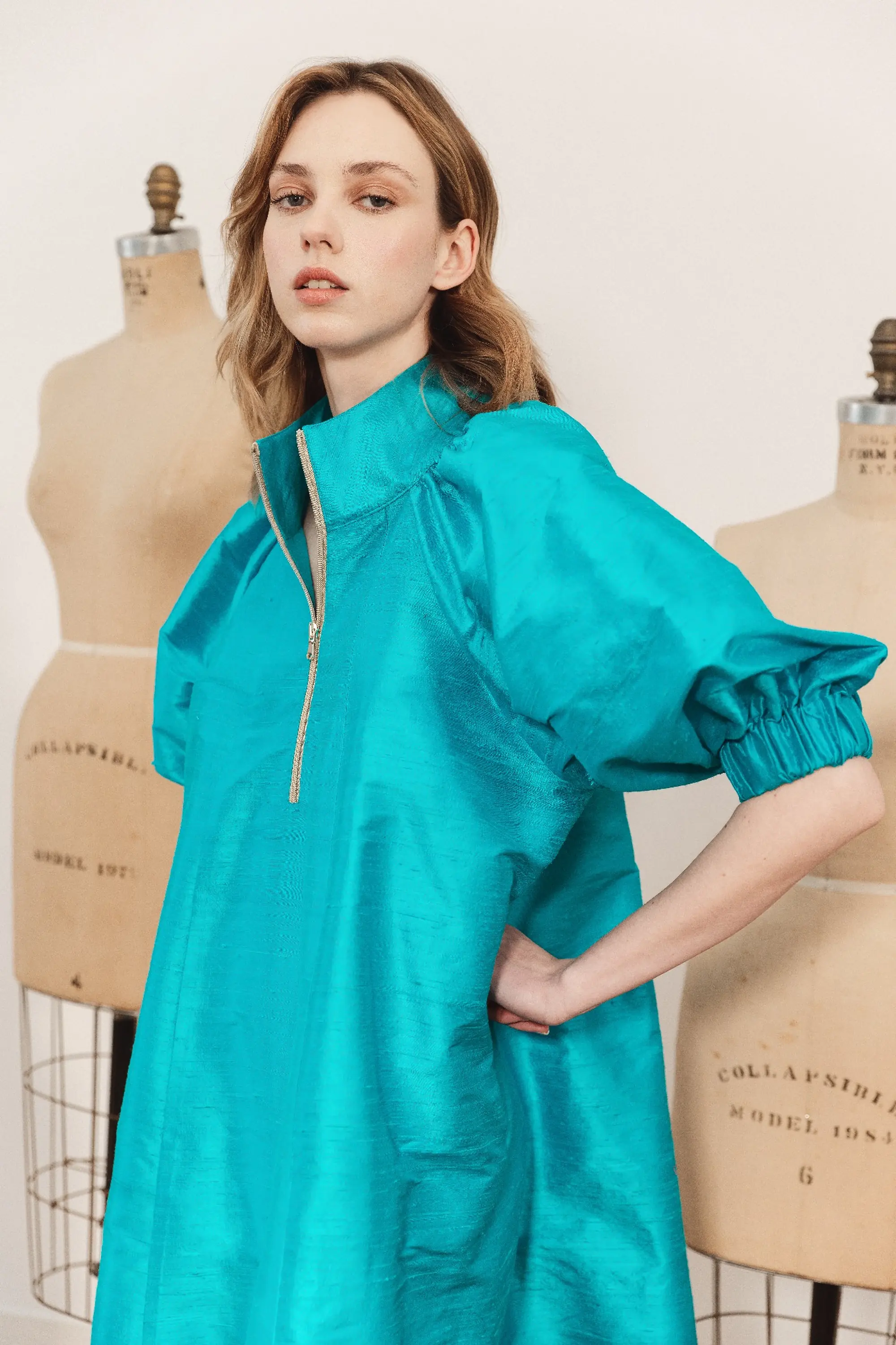 Modella con un abito turchese dalle ampie maniche a sbuffo e zip metallica frontale, fotografata tra manichini sartoriali per la collezione Le Vernissage di André Laug.