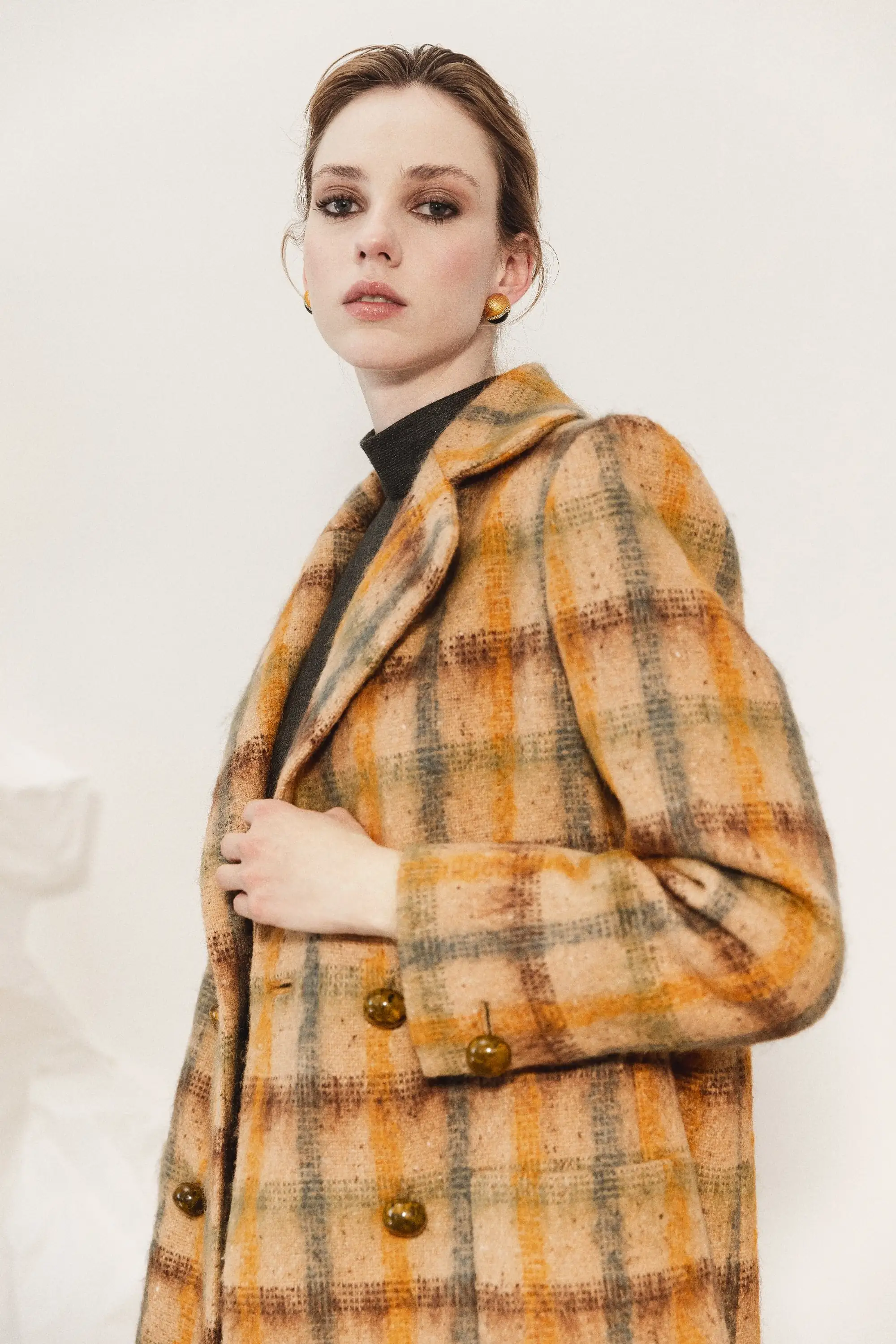 Modella con un cappotto a quadri nei toni del beige, arancio e marrone, indossato sopra un top nero a collo alto, della collezione Le Vernissage di André Laug.