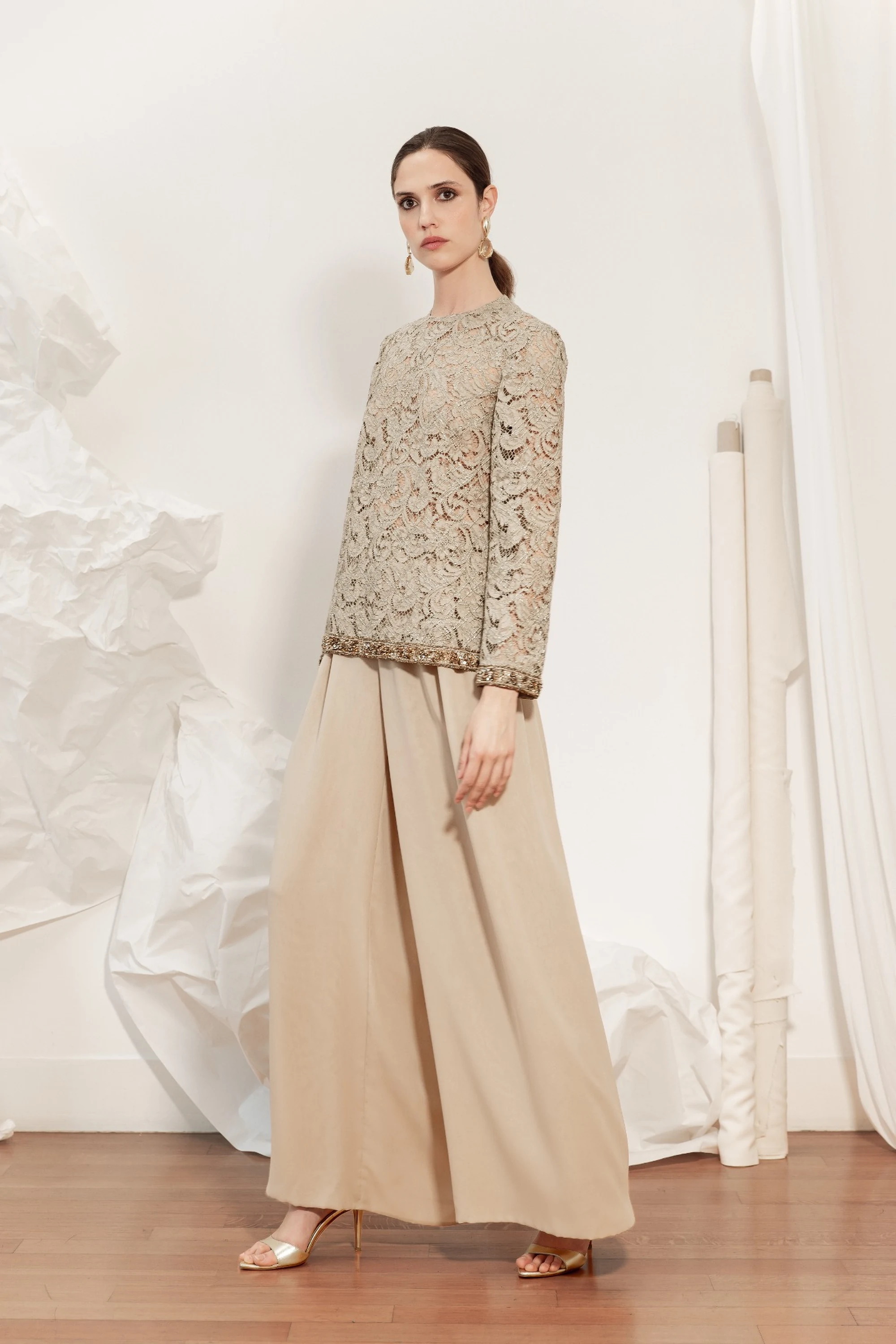 Modella con un top in pizzo color champagne a maniche lunghe, rifinito con dettagli decorativi, e un’ampia gonna lunga beige della collezione Le Vernissage di André Laug.