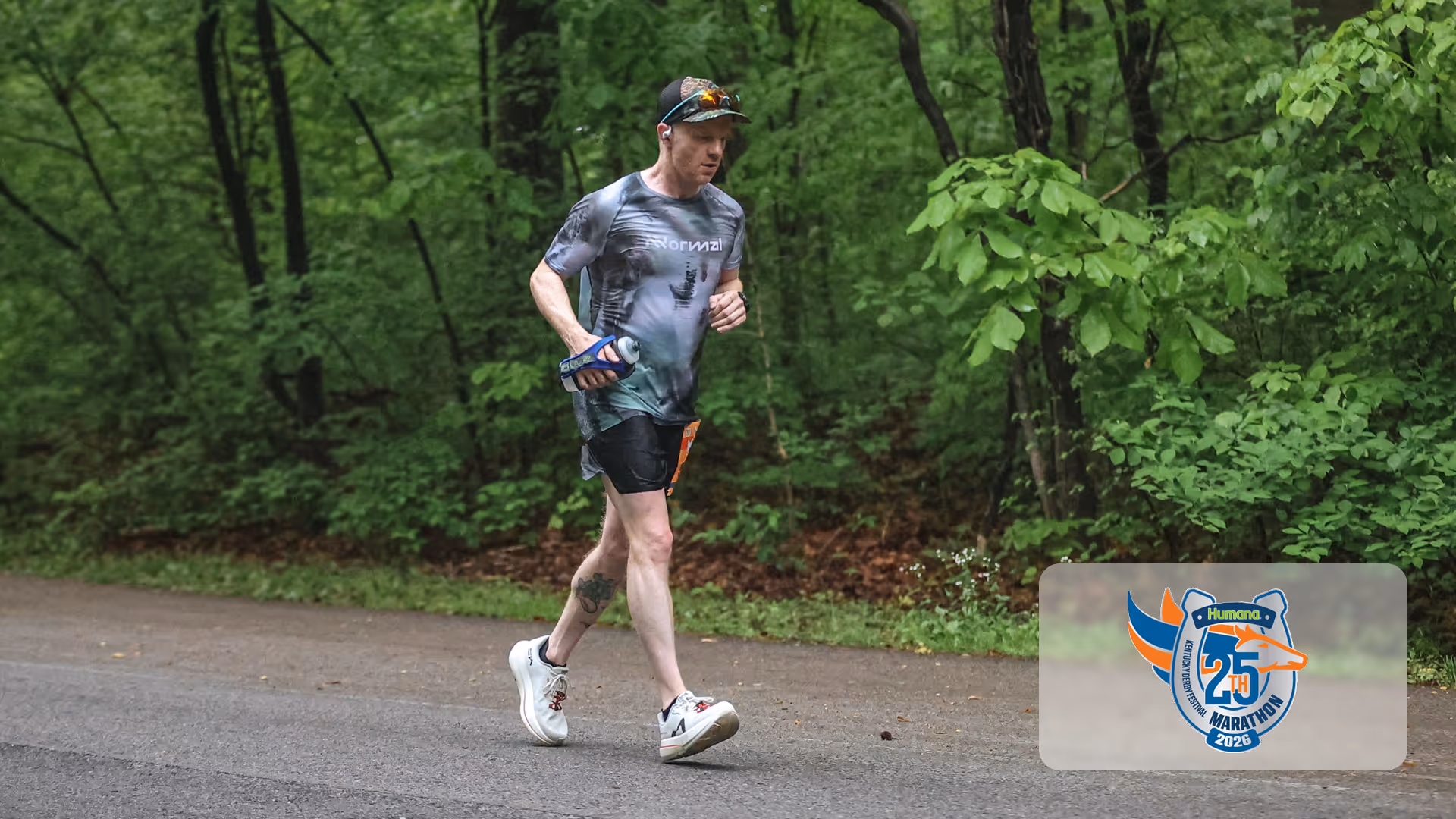 Runner in a gray patterned shirt and black shorts jogging on a road surrounded by green trees.