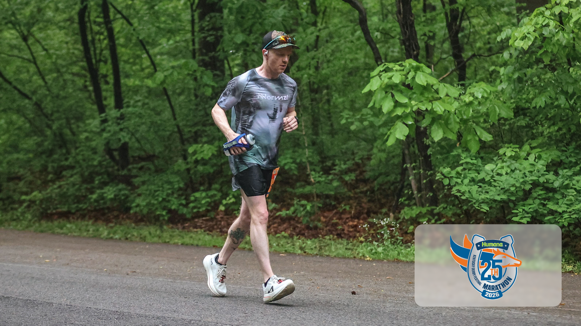 Runner in a gray patterned shirt and black shorts jogging on a road surrounded by green trees.